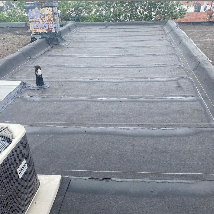 Flat, tar-and-gravel roof with an air conditioning unit and vent pipe. Dark gray surface, overcast day.