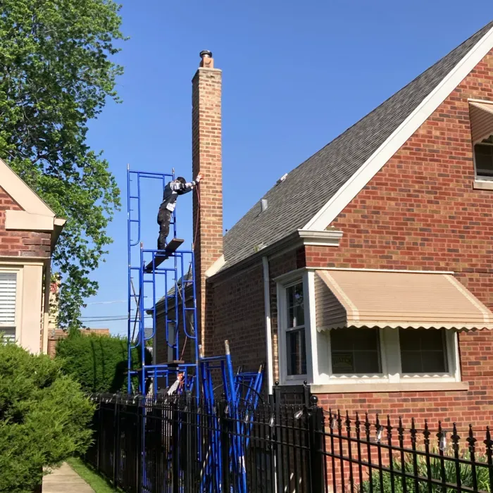 Person on blue scaffolding working on a tall brick chimney attached to a red brick house under a blue sky.
