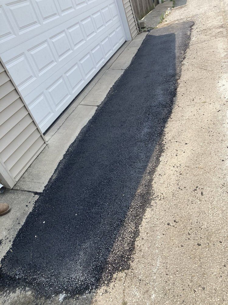 Long, narrow strip of fresh asphalt next to a garage, contrasting with older concrete.