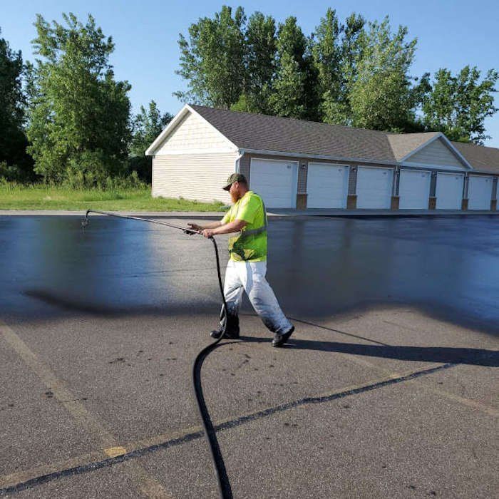 Man in safety gear spraying asphalt sealant on a parking lot, building and trees in the background.
