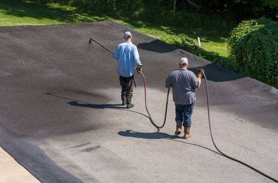 Two workers sealing an asphalt driveway with hoses, dark gray surface.
