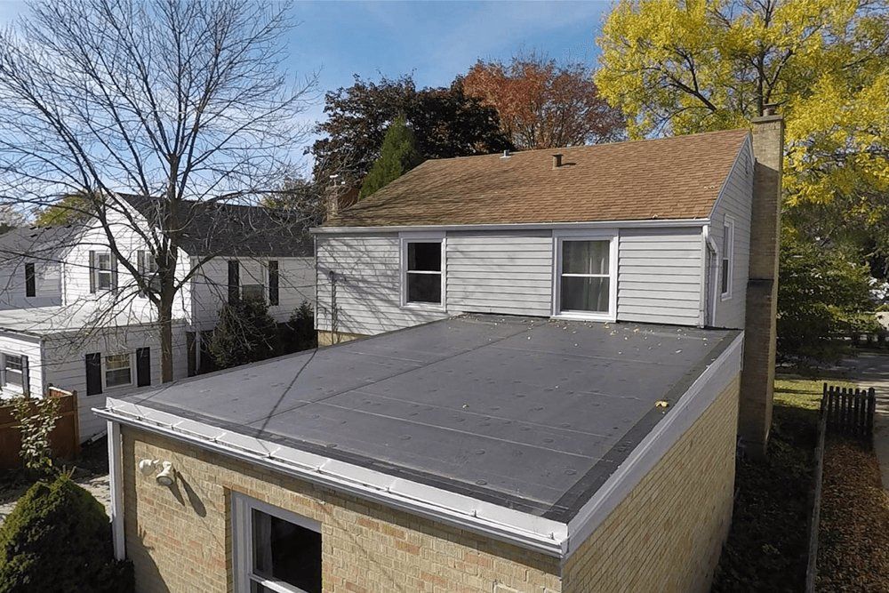 Flat, dark gray metal roof on a light-colored brick building. Two-story home with a brown roof in background.