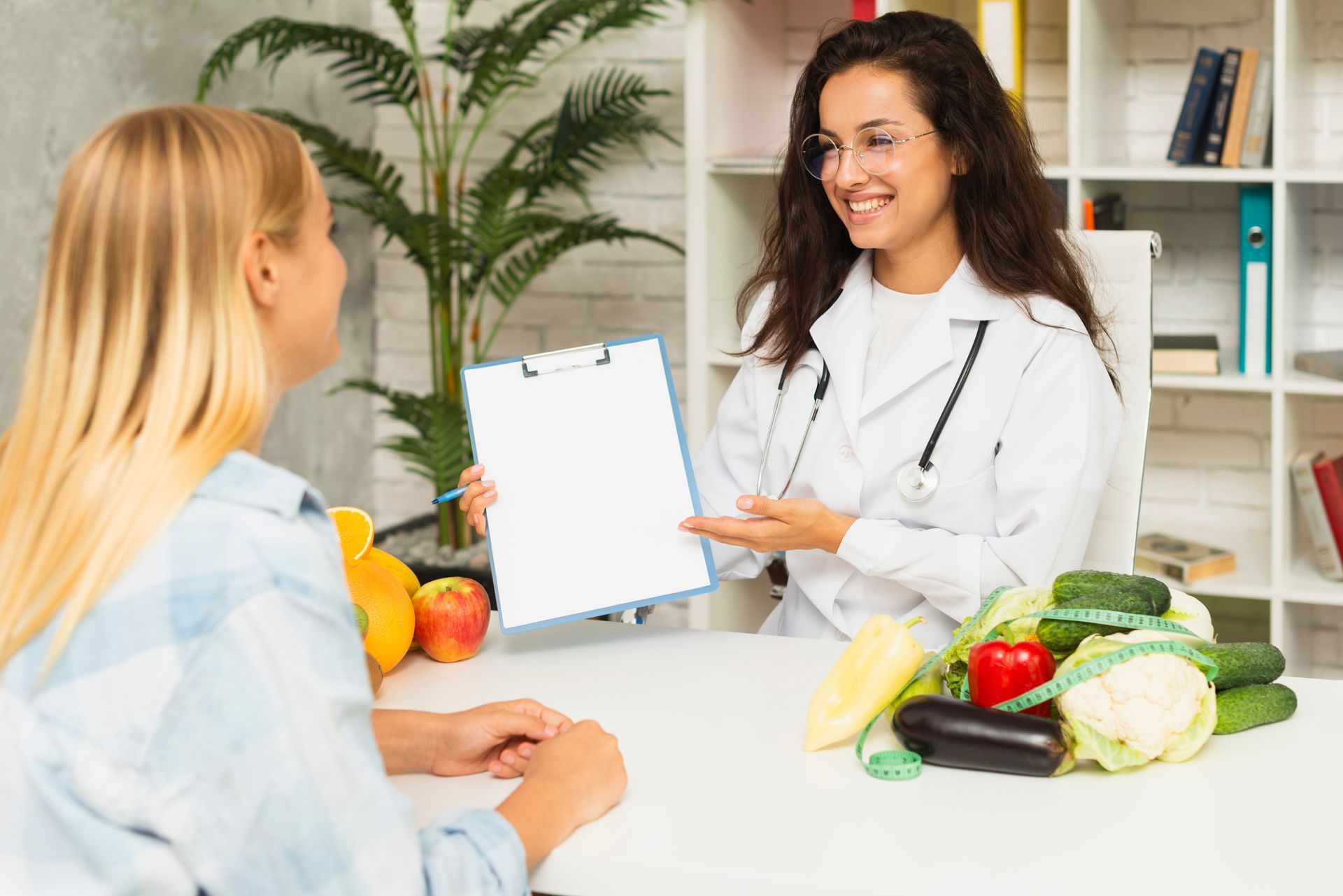 A woman is sitting at a table talking to a doctor who is holding a clipboard.