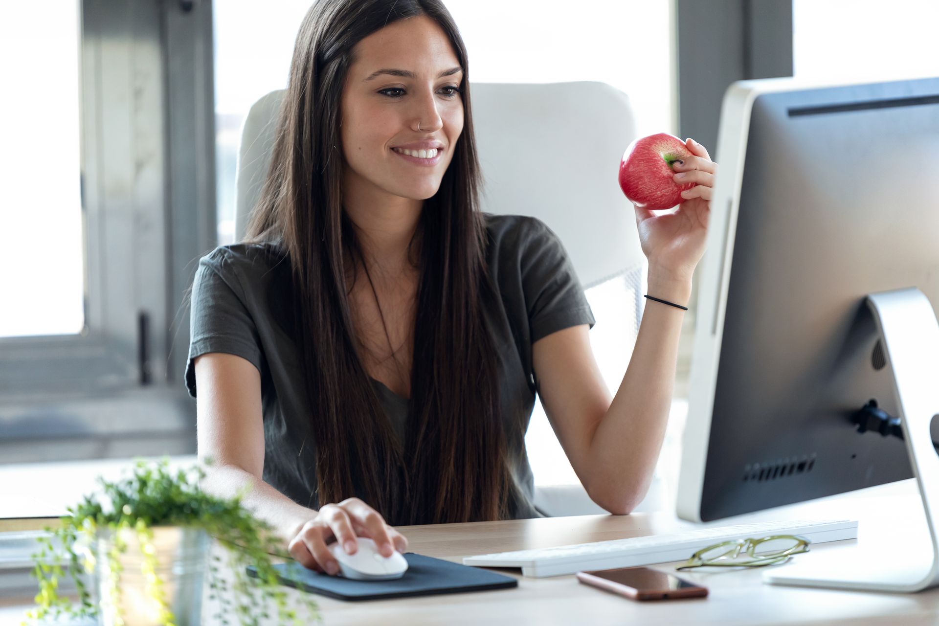 A woman is eating an apple while sitting at a desk in front of a computer.