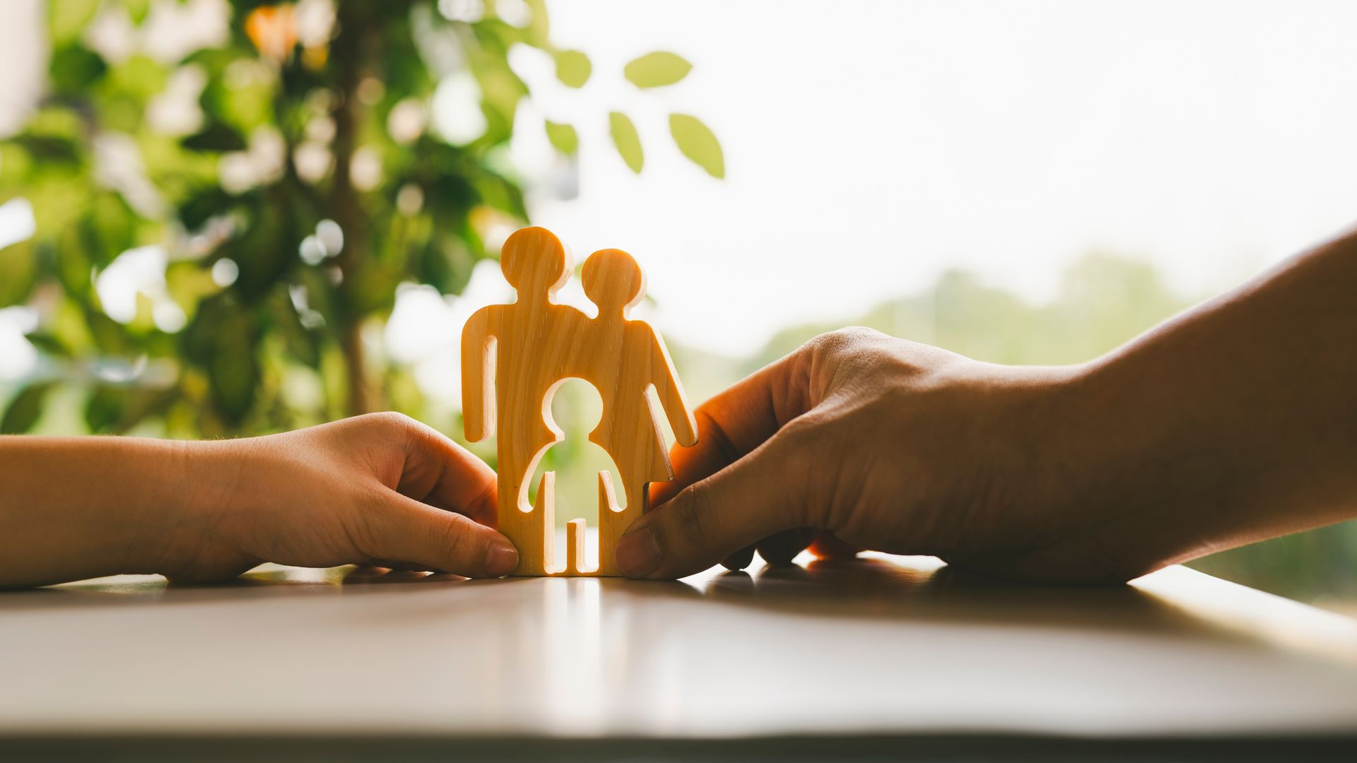 Hands holding a wooden family cutout with a small child figure in front, lit by warm sunlight.