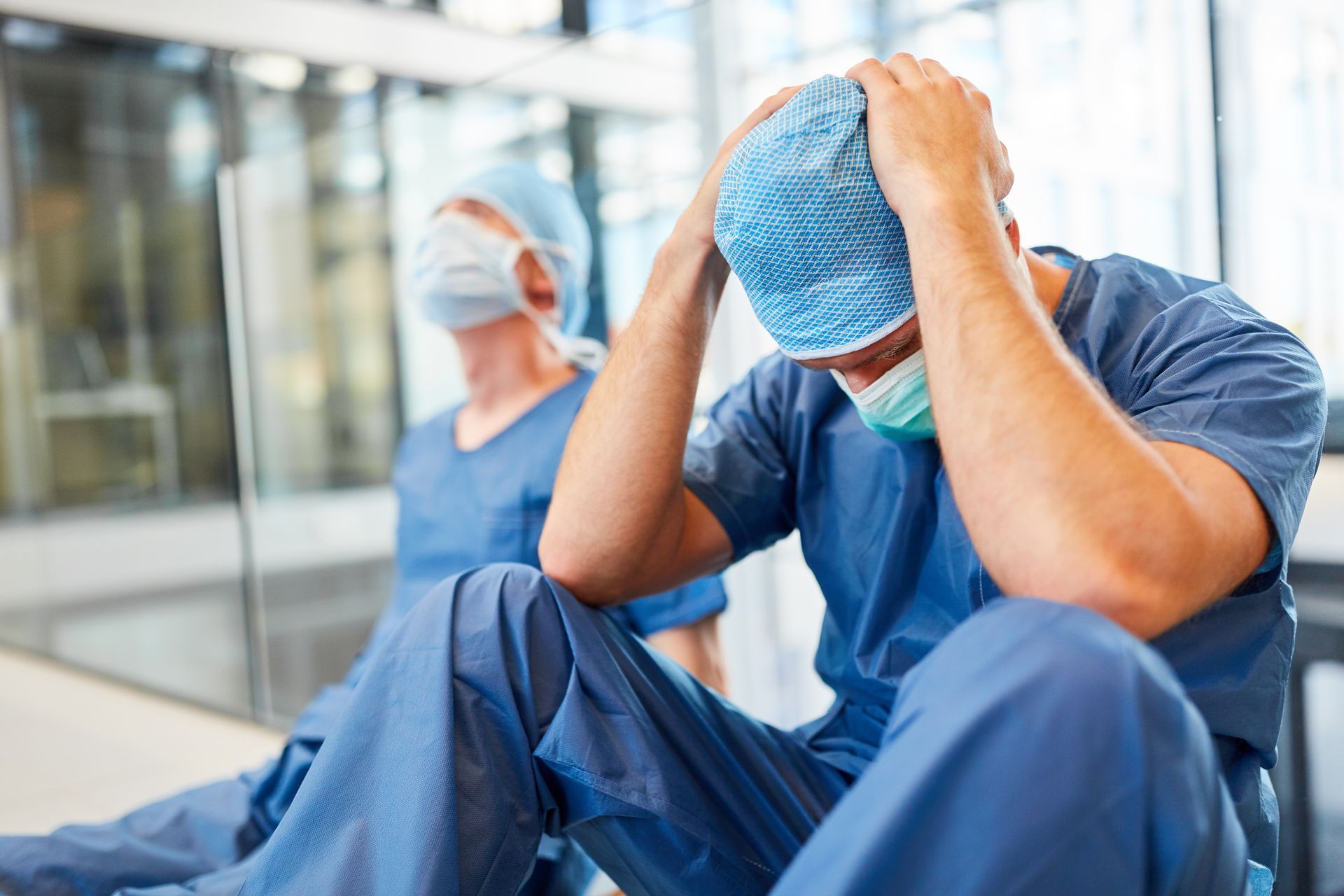 Distressed worker in blue scrubs sitting on the floor, holding head, with a masked coworker in the background