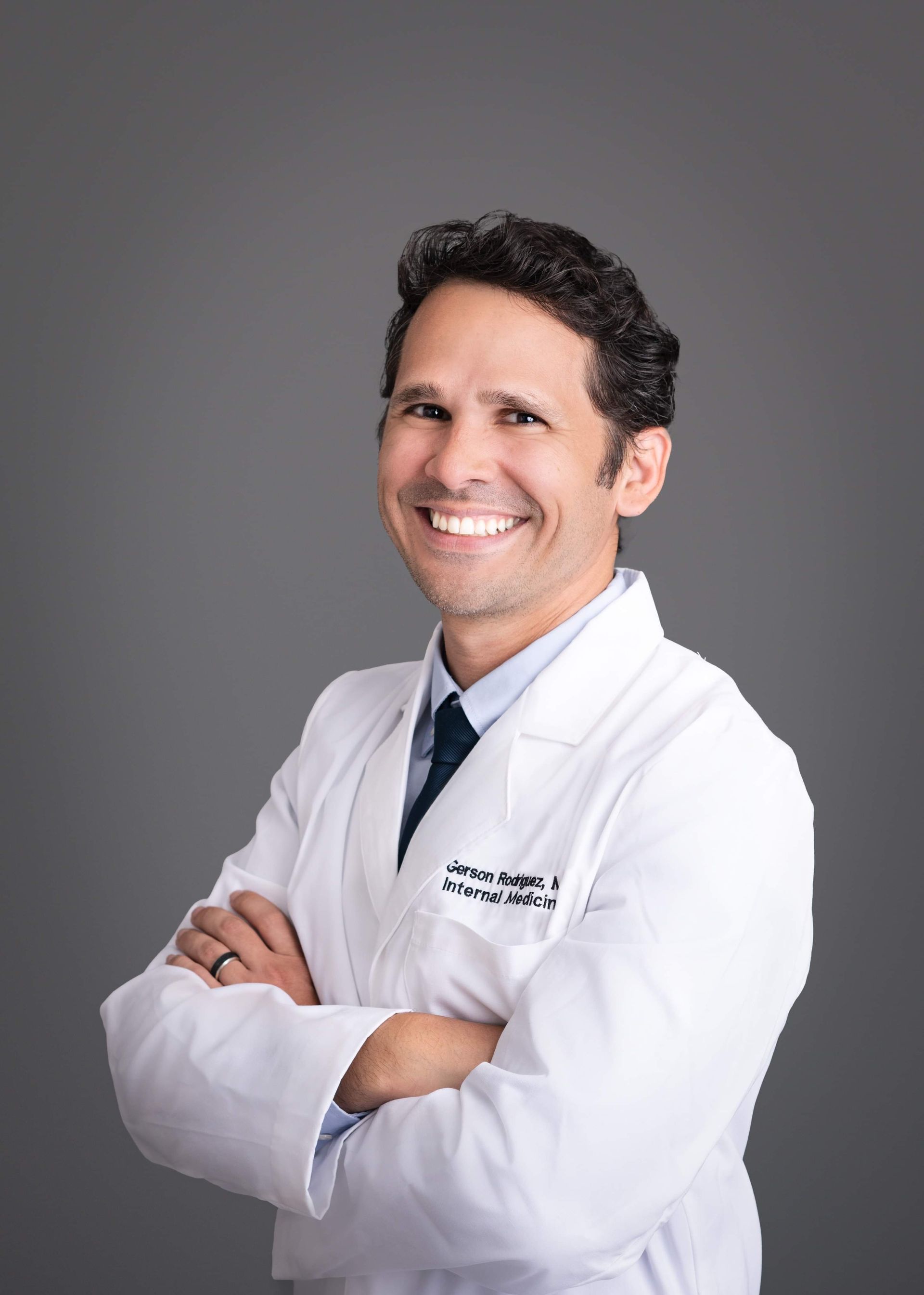 A man in a white lab coat and tie is smiling with his arms crossed.