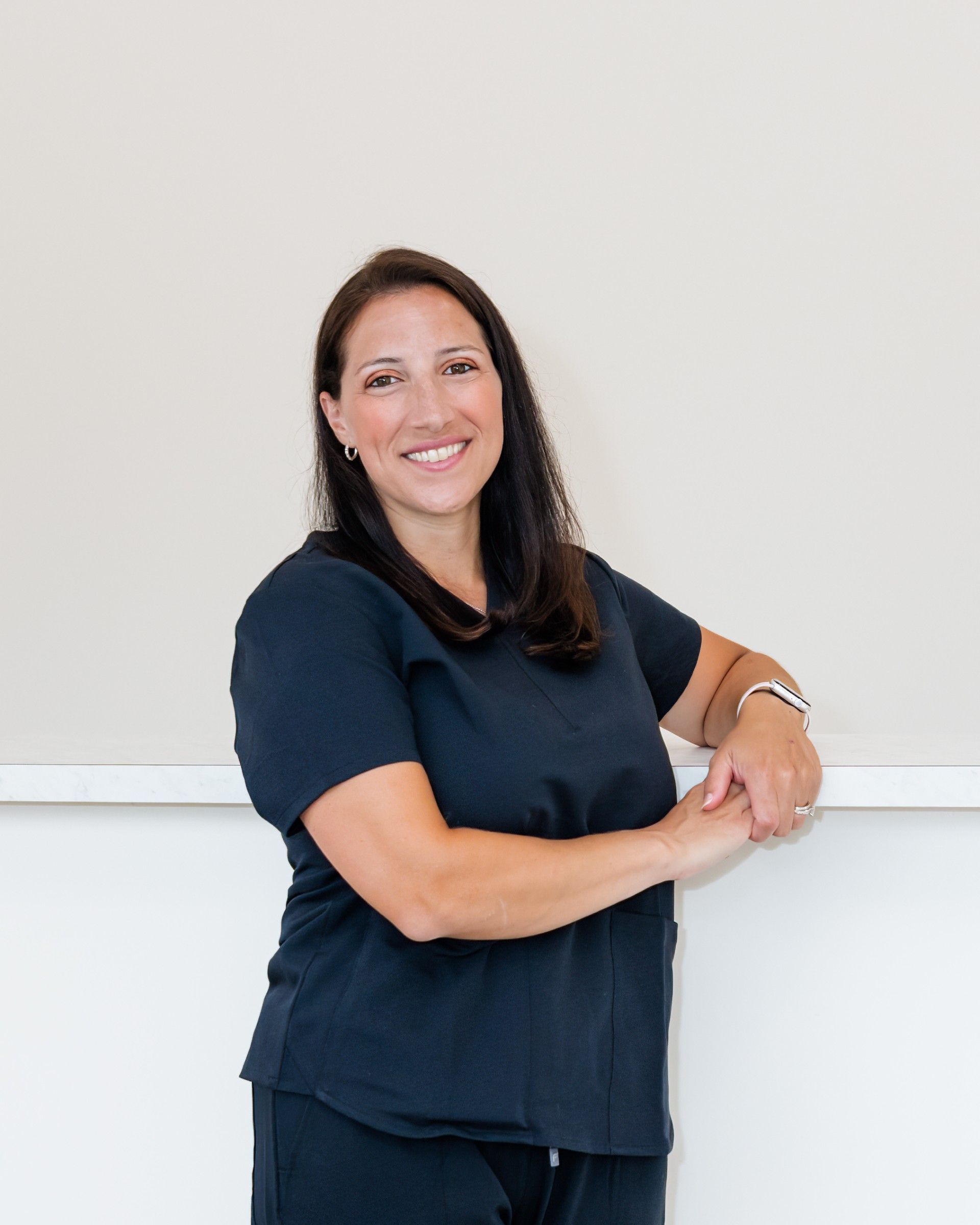 A woman in a black scrub top is leaning against a wall and smiling.