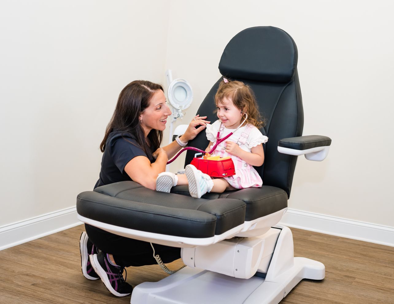 A woman is kneeling down next to a little girl sitting in a chair.
