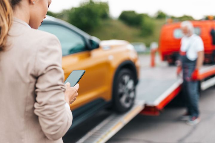 Person with phone beside a car on a tow truck, with another person in the background.