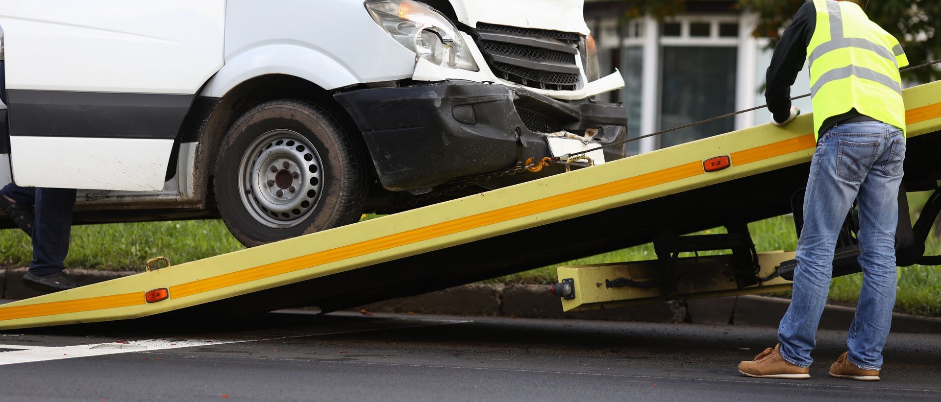 Tow truck loading a damaged white van, with a worker in a reflective vest guiding the ramp.