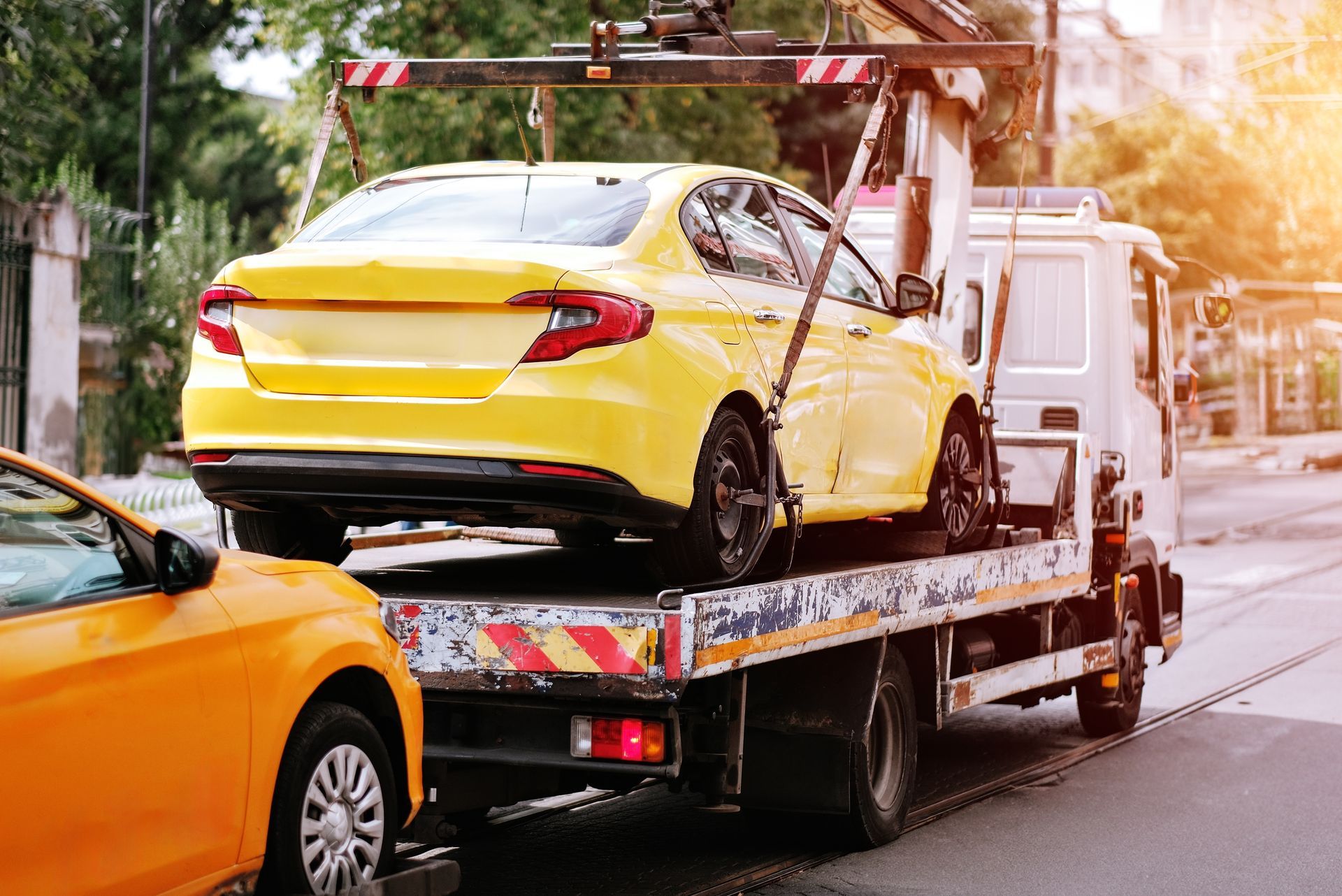 Yellow car being towed on a flatbed truck beside an orange car on a city street.