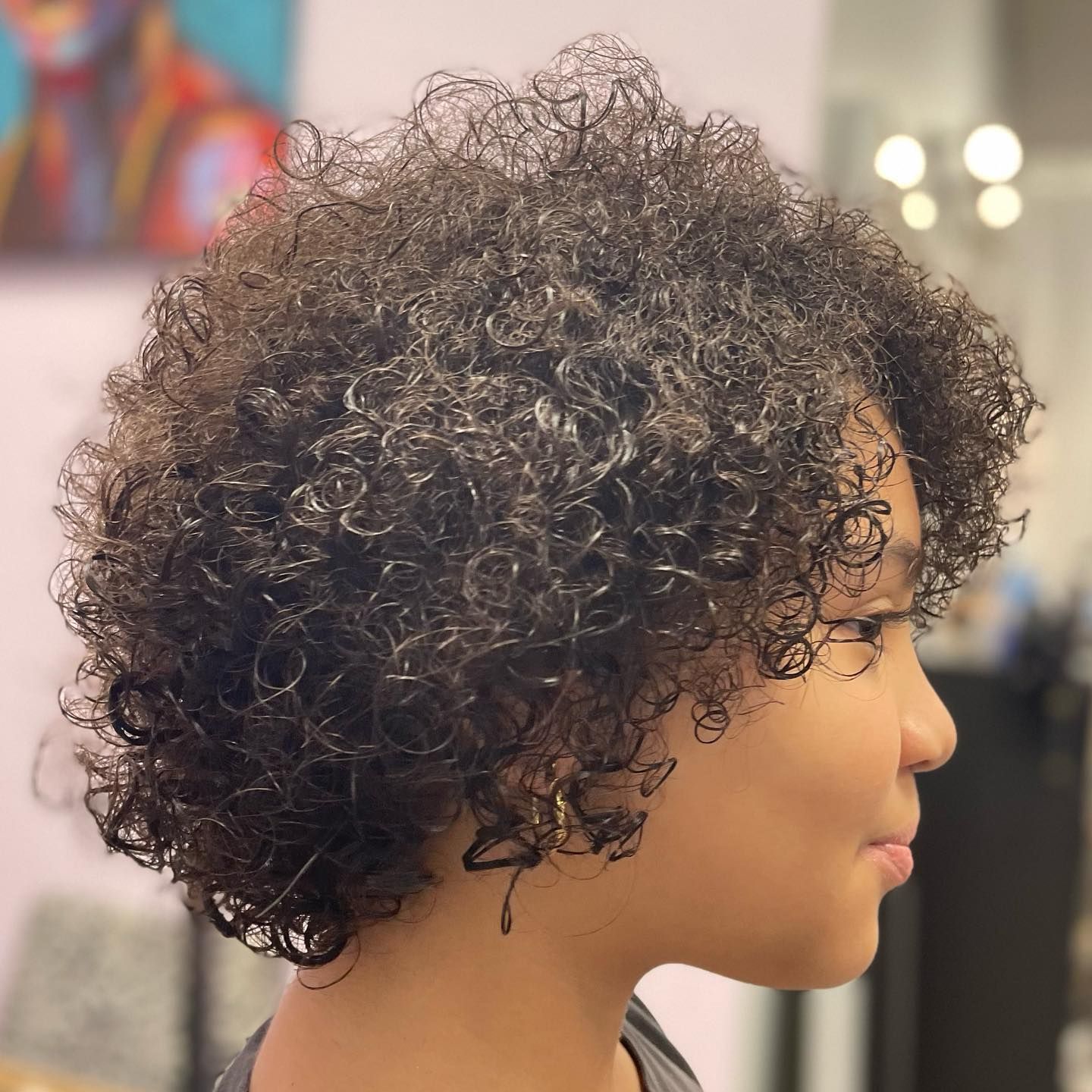 Person with curly dark hair, side profile. Inside, with artwork and lights in background.