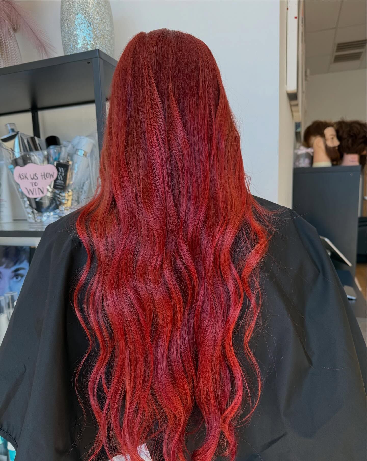 Person with long, wavy, bright red hair in a salon, wearing a black cape.