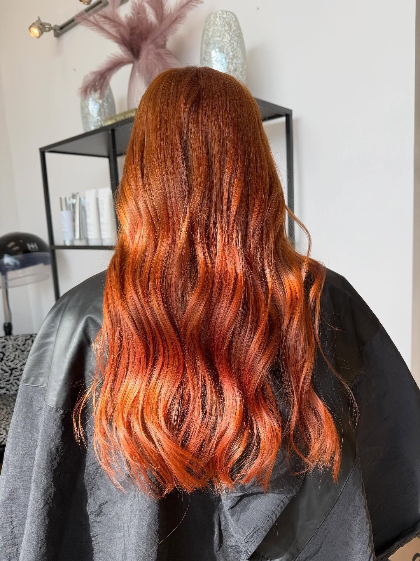 Woman with long, wavy auburn hair. A black cape is around her shoulders. In a salon.