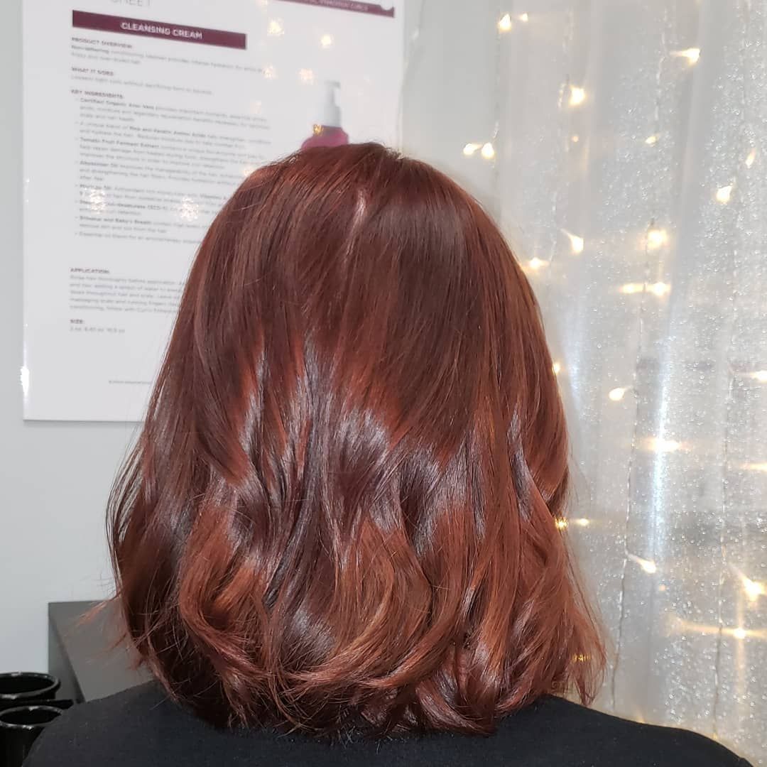 Woman with reddish-brown wavy hair, viewed from behind. Light reflects on the glossy color.