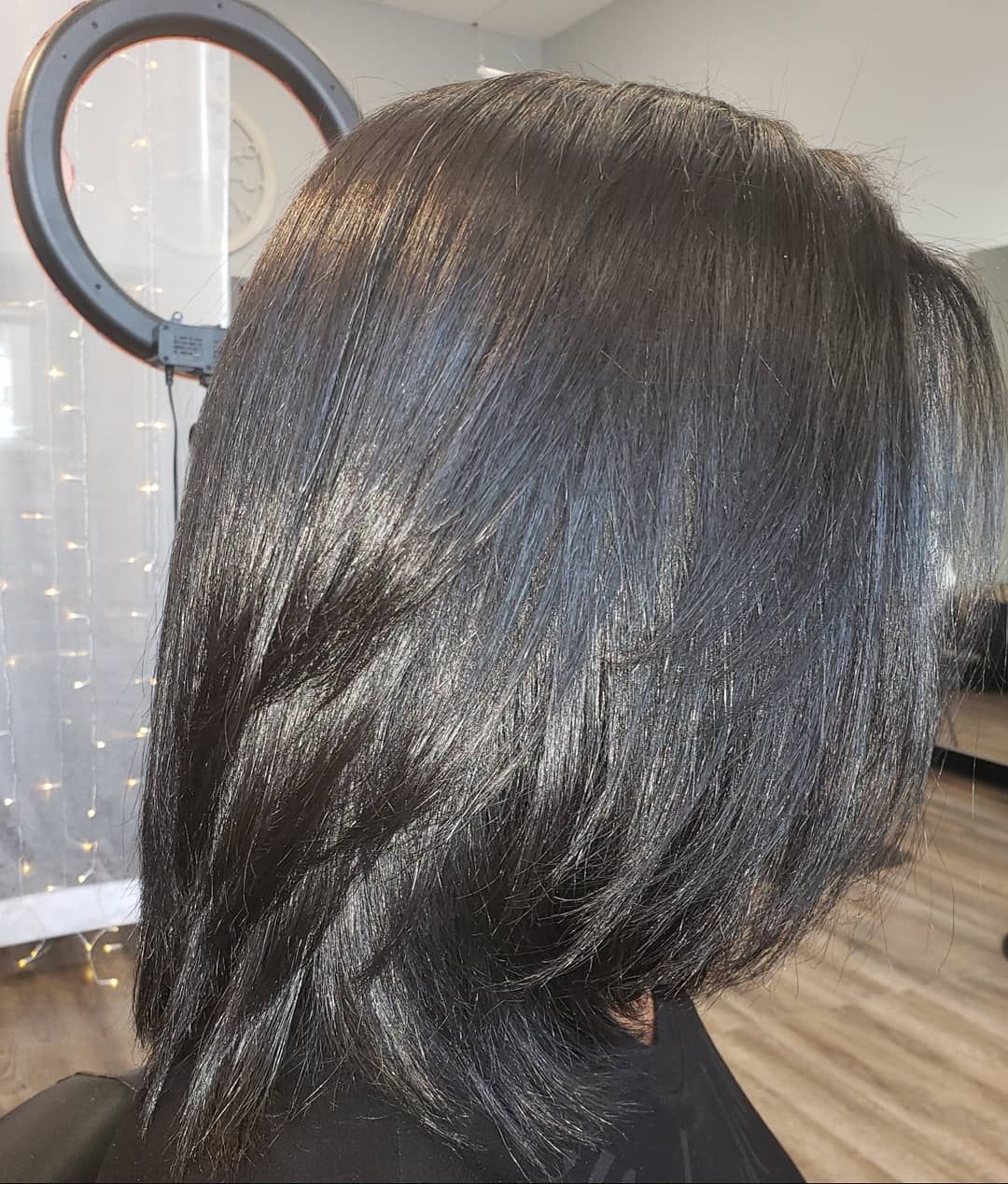 A person with sleek, dark, layered hair in a salon setting, lit by a ring light.