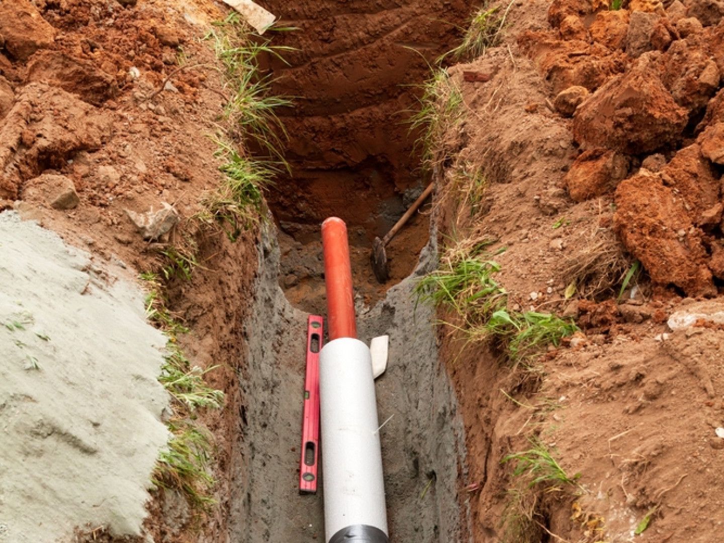 Trench with a white pipe and a red conduit inside; a level rests against the pipe.