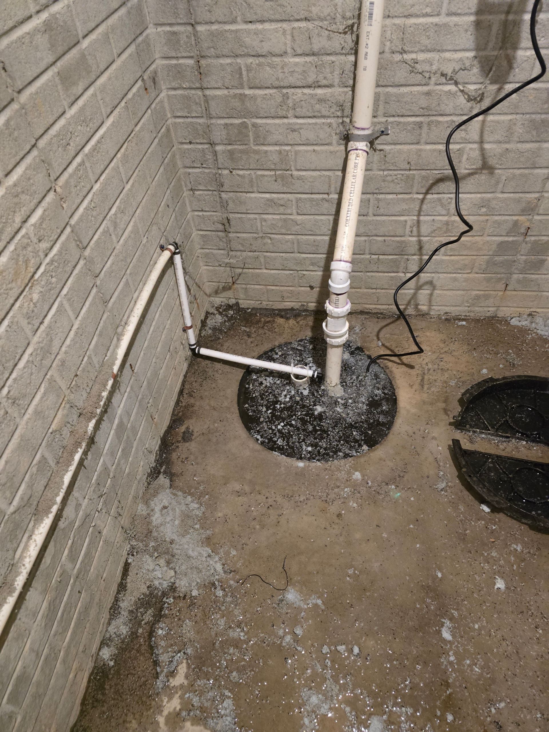A basement sump pump installation, with white PVC piping against a brick wall and a dark cover.
