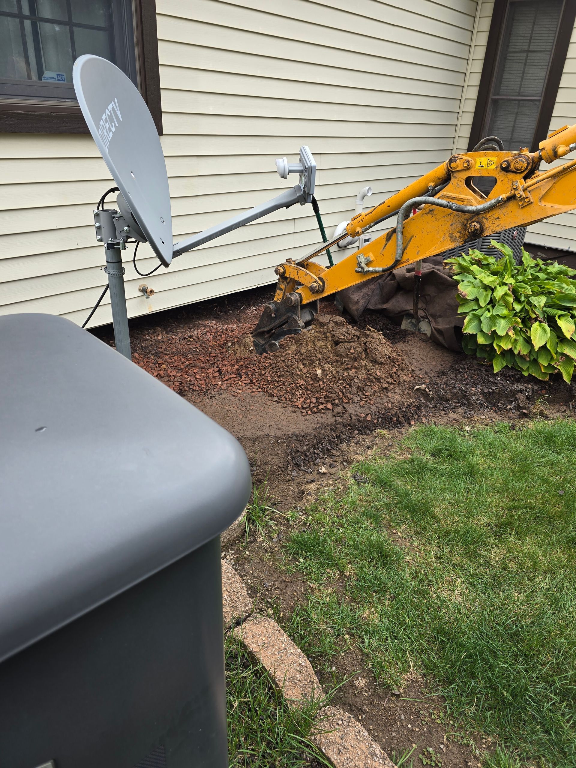 Yellow excavator digging in a residential yard near a satellite dish and shrub.