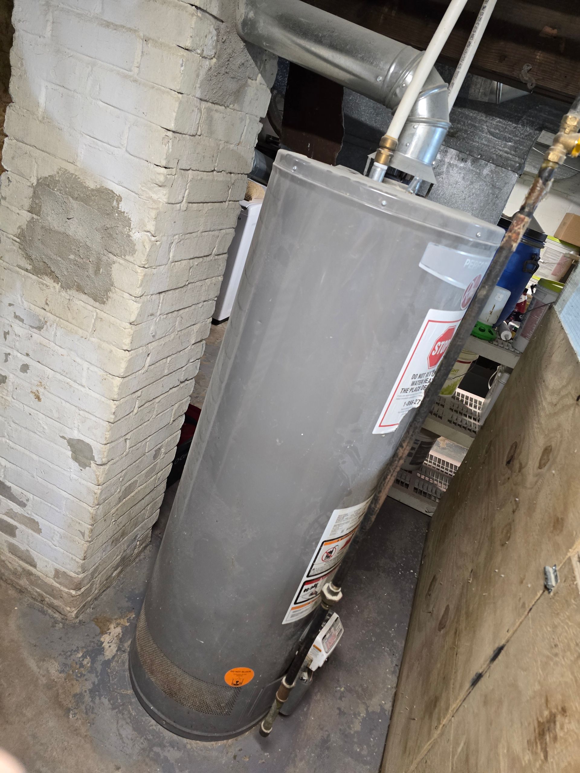 Water heater in a basement. The appliance is gray and tall, positioned near a brick wall and wooden paneling.