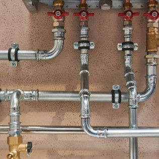 Pipes, valves, and fittings in a plumbing system, mostly silver with red and brass accents, against a beige wall.