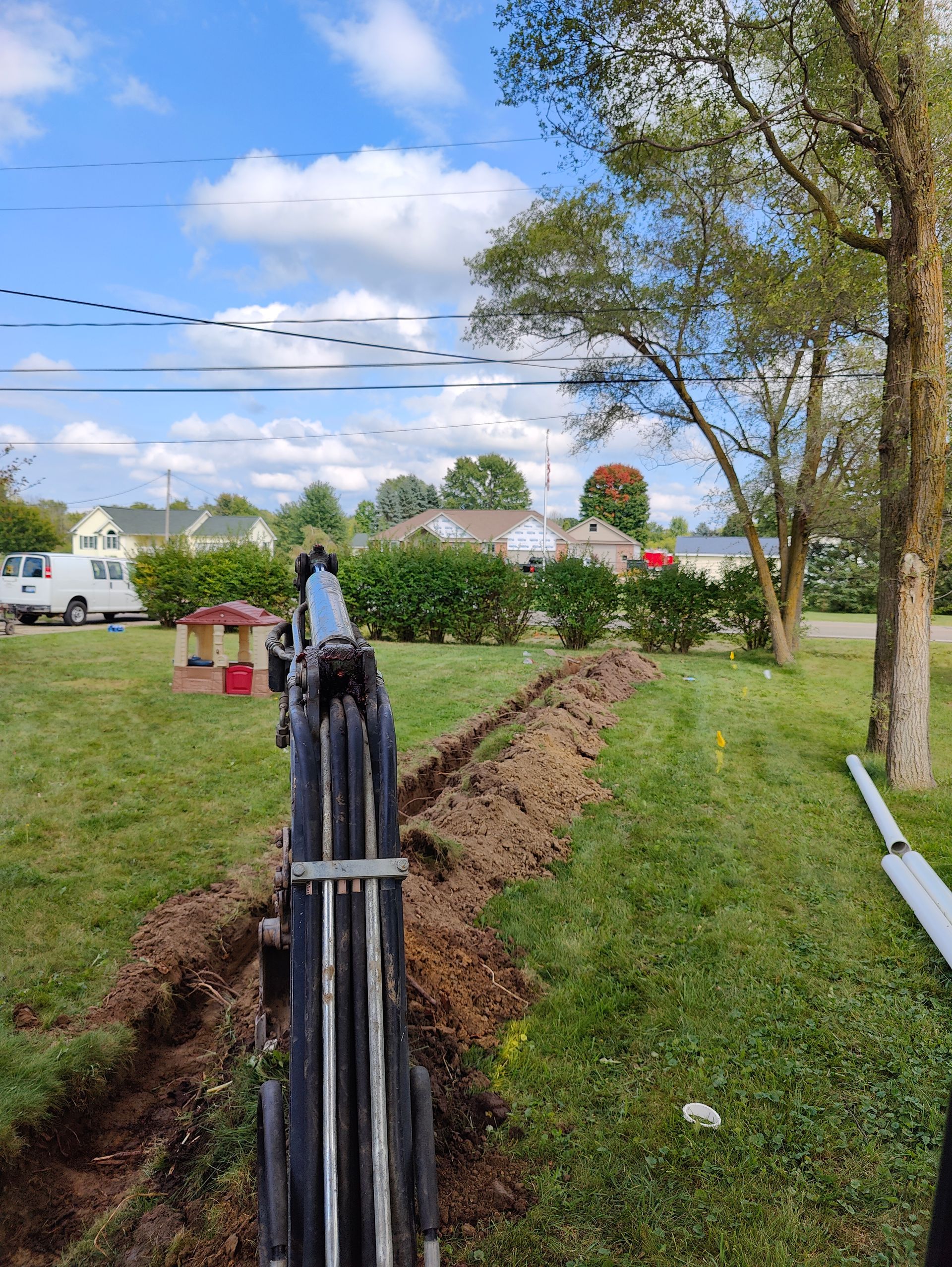 A backhoe digging a trench in a grassy yard near a tree and houses under a blue sky.