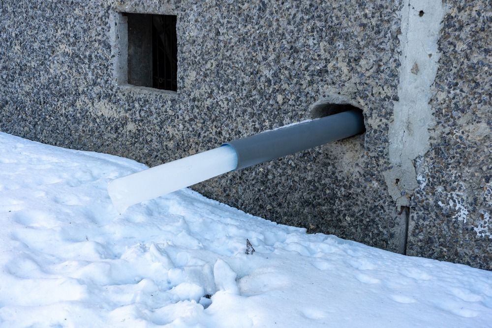 Gray pipe, partially ice-covered, exits a concrete wall. Snow covers the ground.
