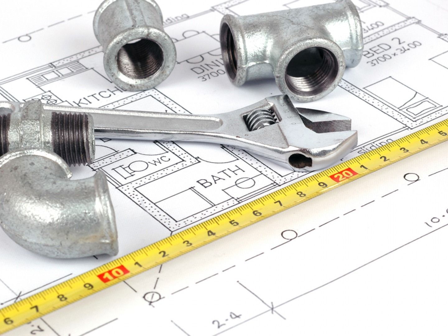 Plumbing tools and fittings on top of a blueprint with a measuring tape and adjustable wrench.