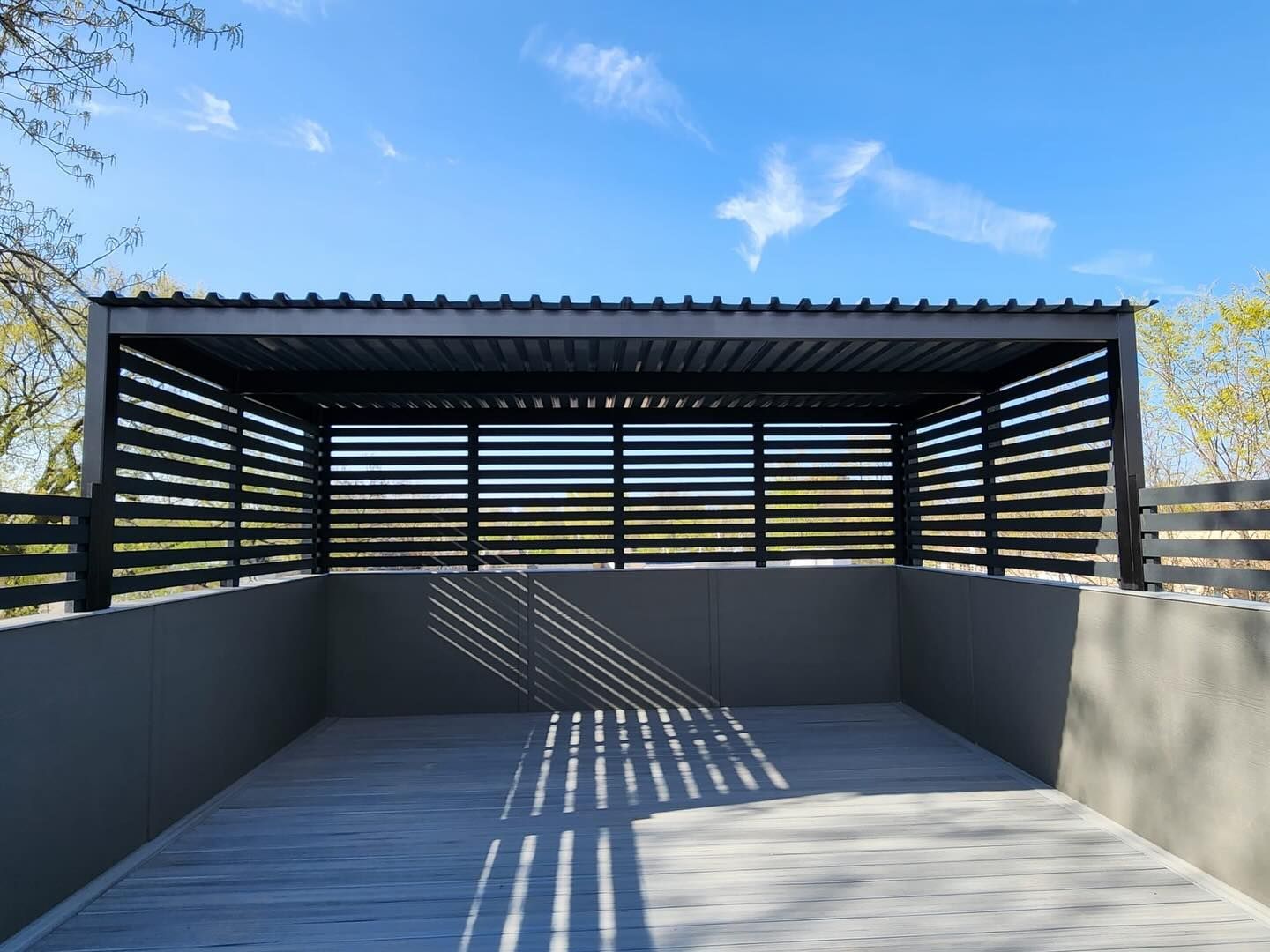A modern, dark-roofed pergola with horizontal slats casting shadows on a concrete patio, against a blue sky.