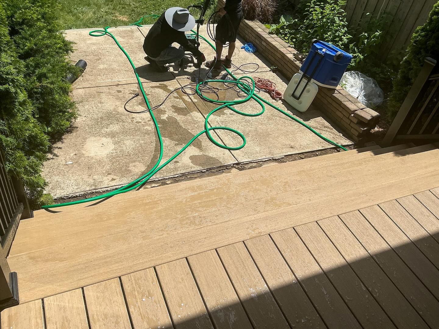 Two people repairing a concrete patio. Green hose outlines the area. Deck in foreground.