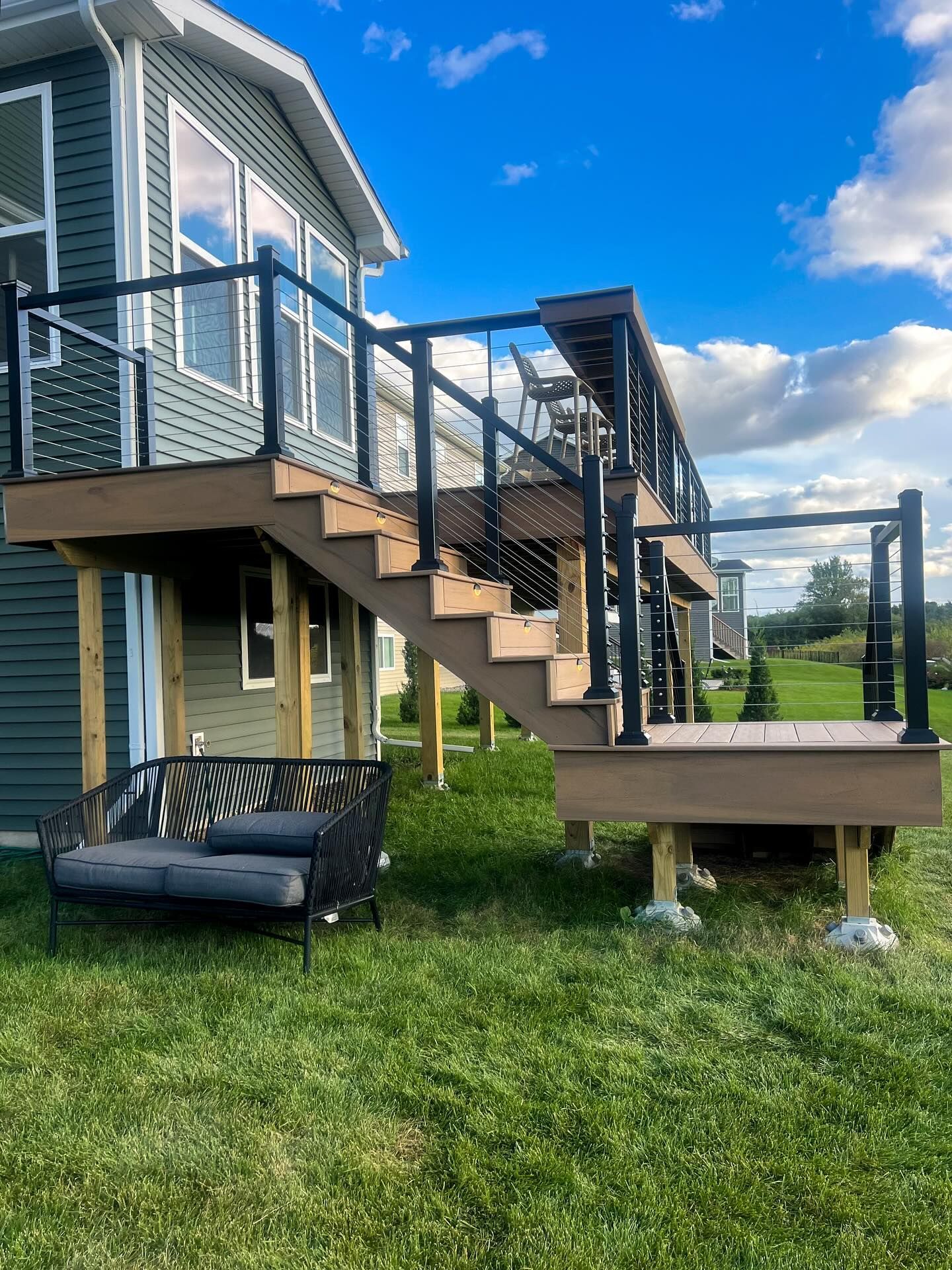 Green building with a brown deck, stairs, black railings, and a gray outdoor sofa on green grass under a blue sky.