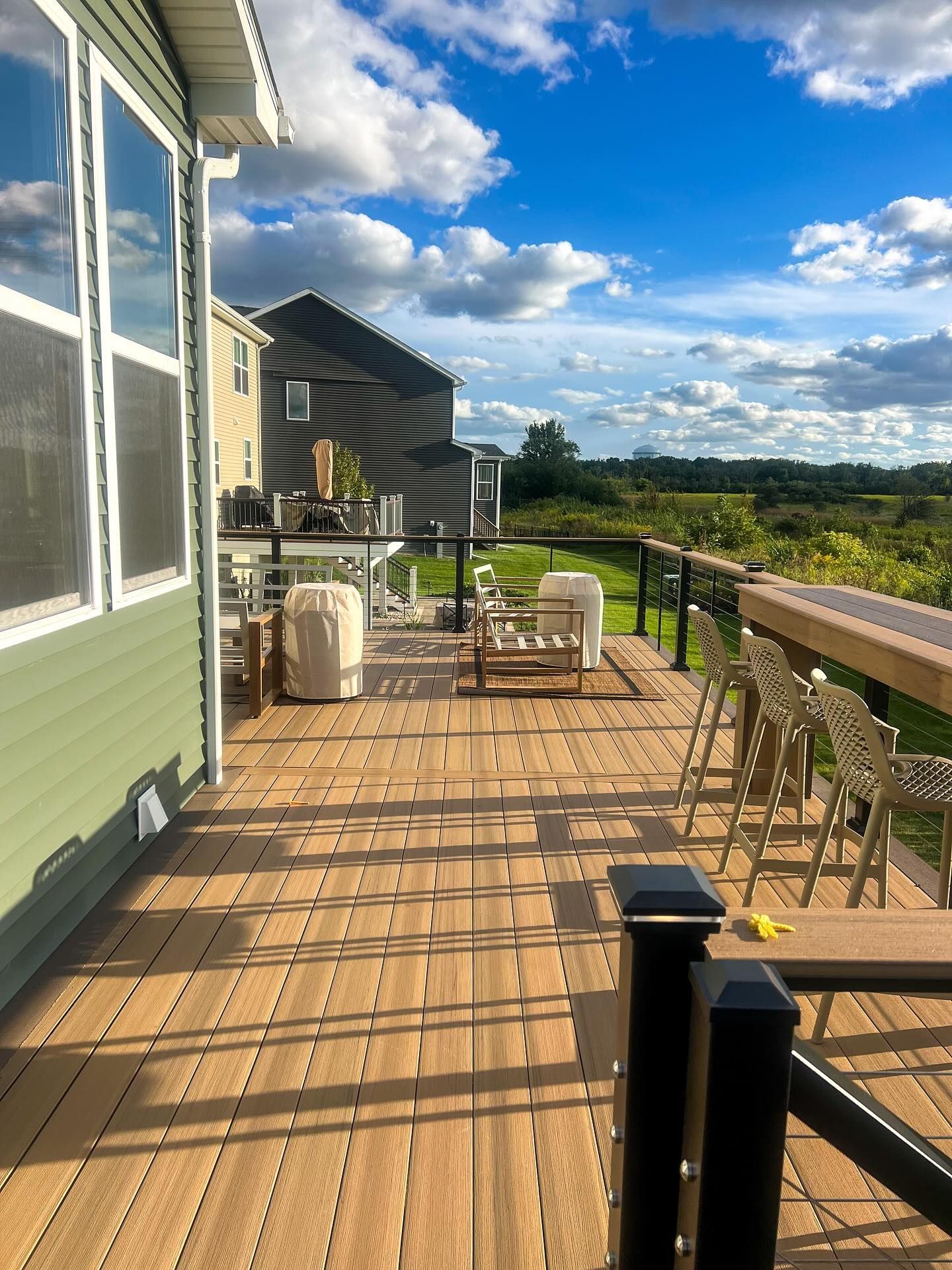 Wooden deck with seating and railing overlooking a green field under a blue sky.