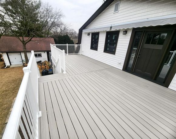 A gray wooden deck with white railings extends from a white house with dark doors and windows.
