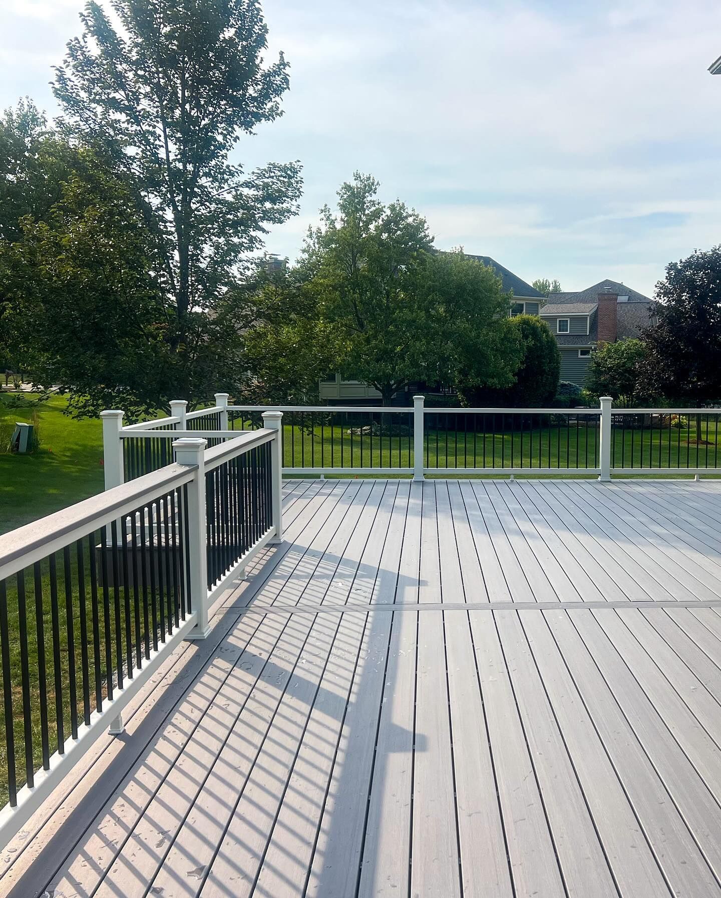 Grey wooden deck with white railing, black spindles, and trees in background under a sunny sky.
