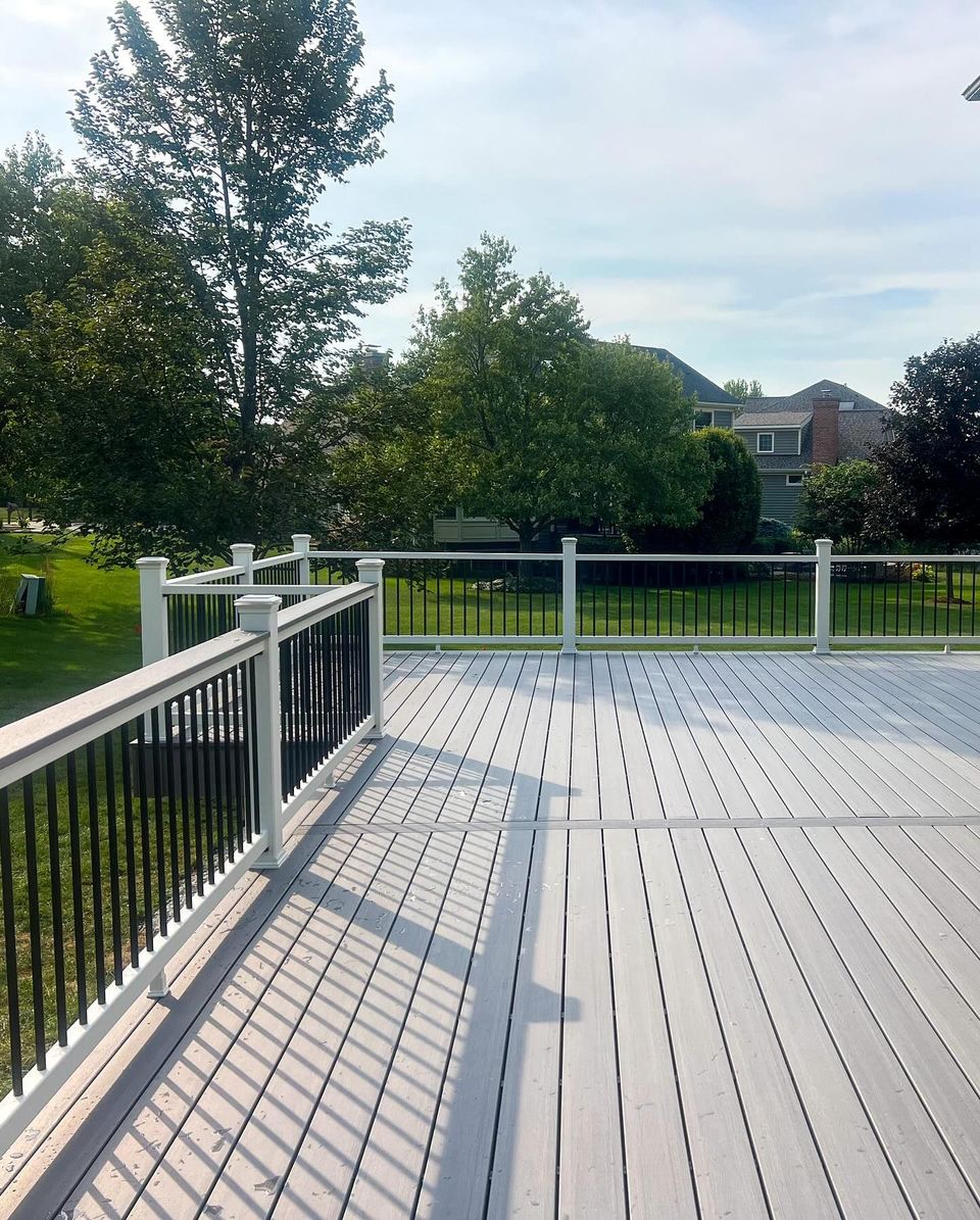 Grey wooden deck with white railing, black spindles, and trees in background under a sunny sky.