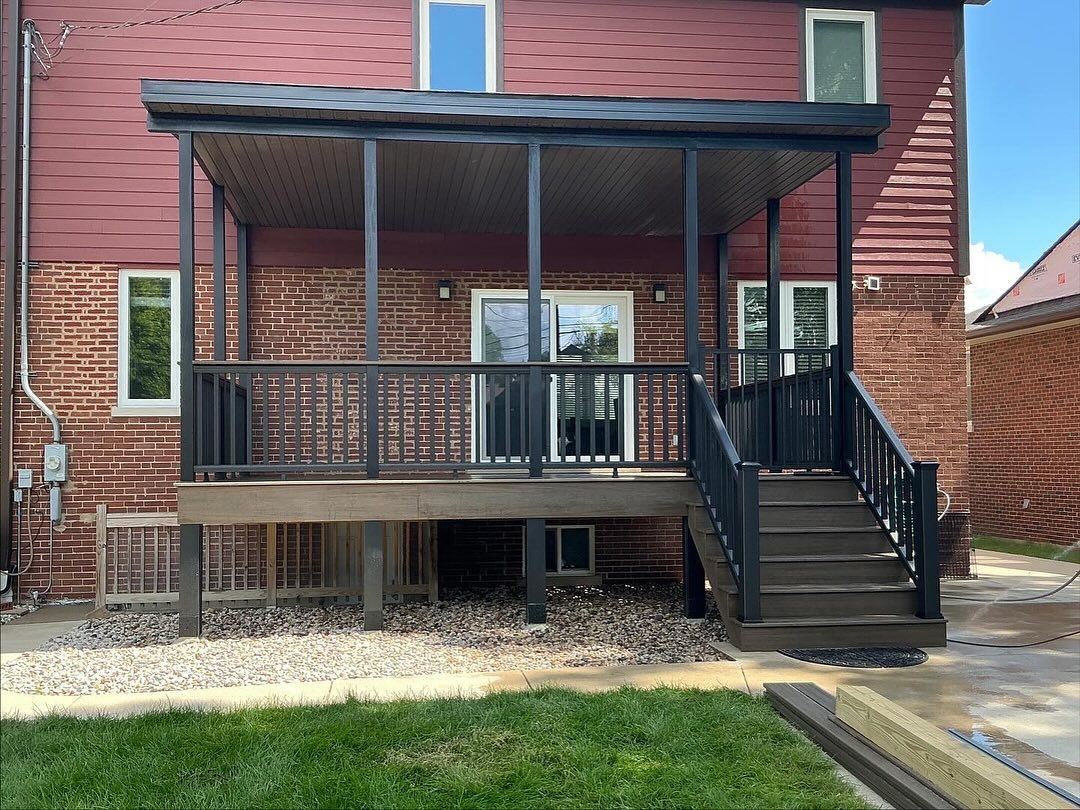 Covered back porch with black railings, stairs, and support posts attached to a red brick and wood-sided house.