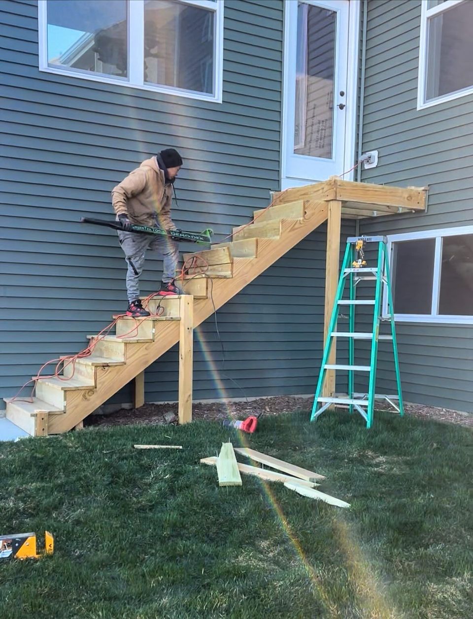 Man building wooden stairs attached to a house. Green ladder next to the stairs. Outdoors, daylight.