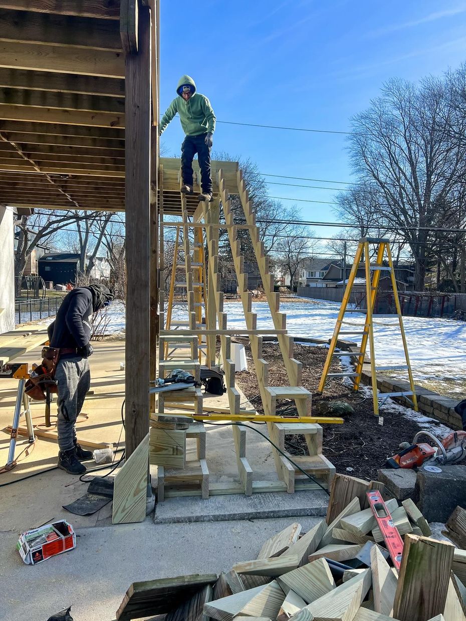 Construction of wooden stairs leading to a deck; two people working. Snowy ground, sunny day.