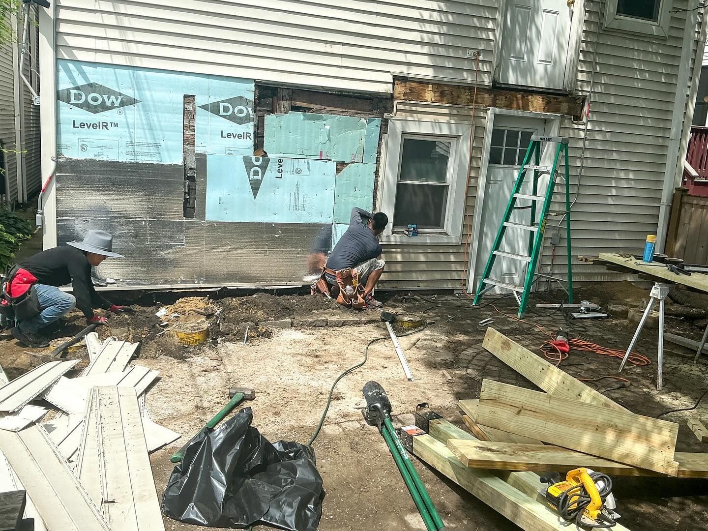 Construction workers repairing exterior wall of a house. Scaffolding, wood, and insulation are visible.