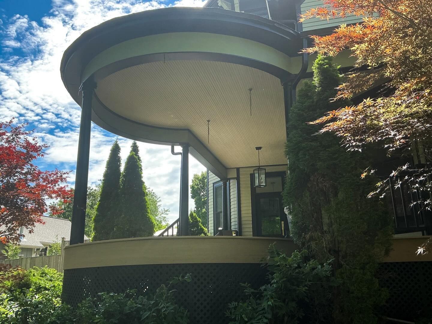 Curved porch of a house with black columns, green trim, and trees. Sunny day.