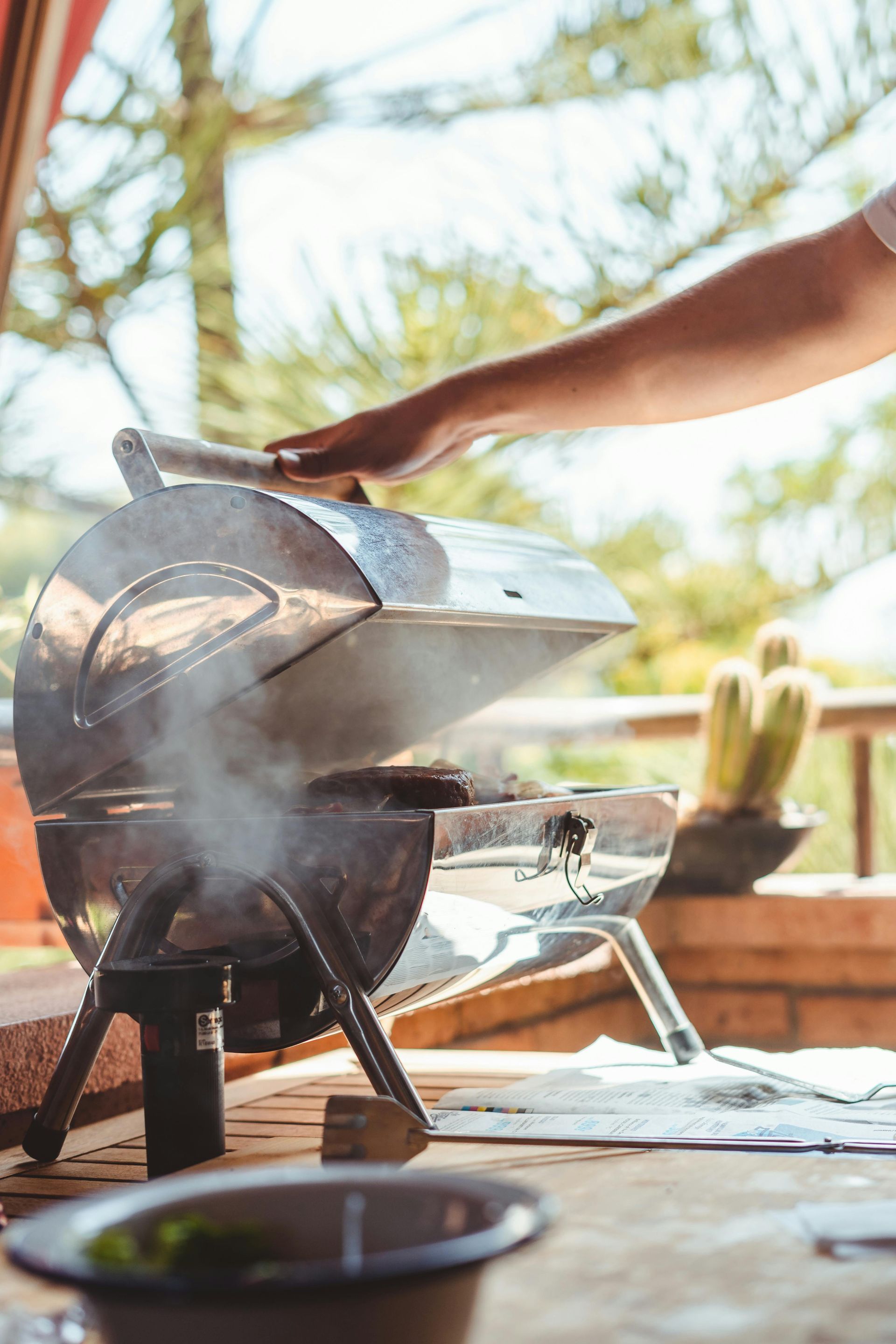 Person grilling on a small outdoor barbecue, hand on lid, steam rising.