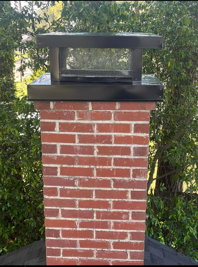 A brick chimney with a black top is surrounded by trees.