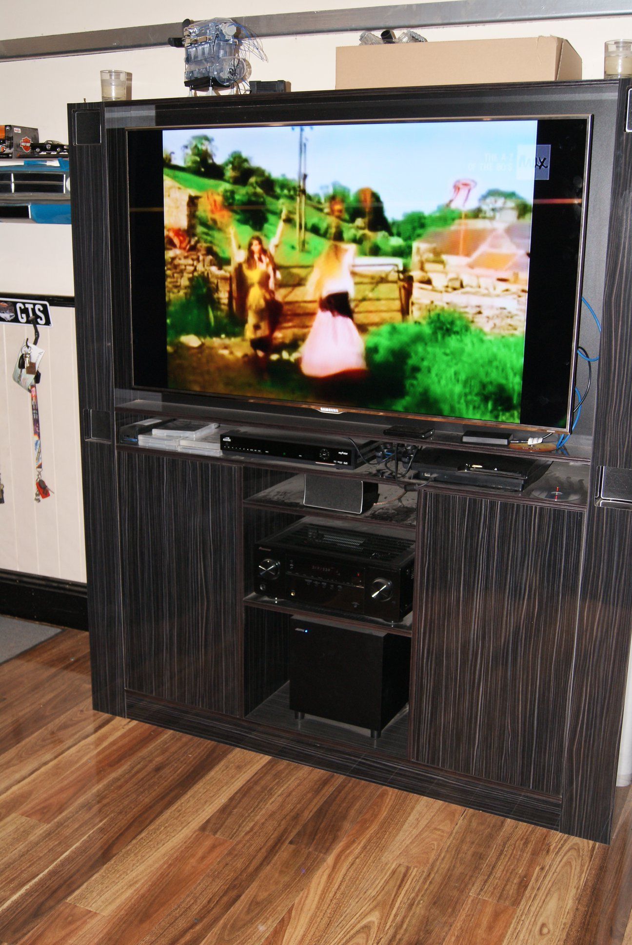 TV cabinet with a TV playing a scene outdoors; the cabinet is black with a wood grain finish and houses other electronic devices.