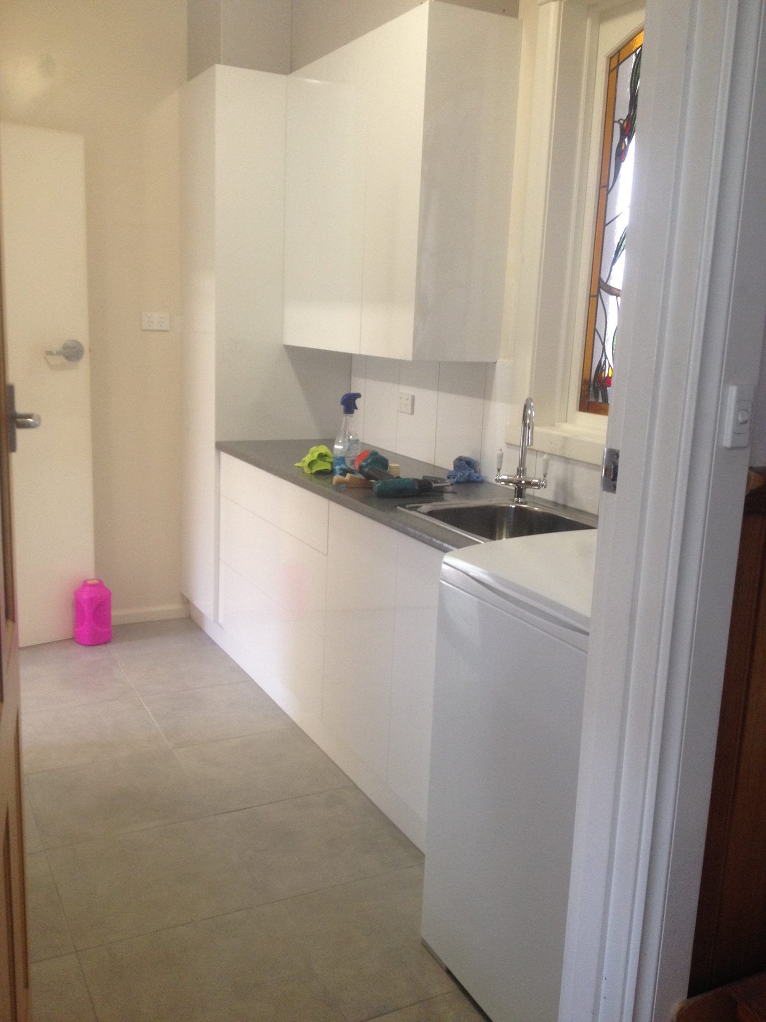 A laundry room with white cabinets, a washing machine, and a sink. The room has a window and concrete floor.