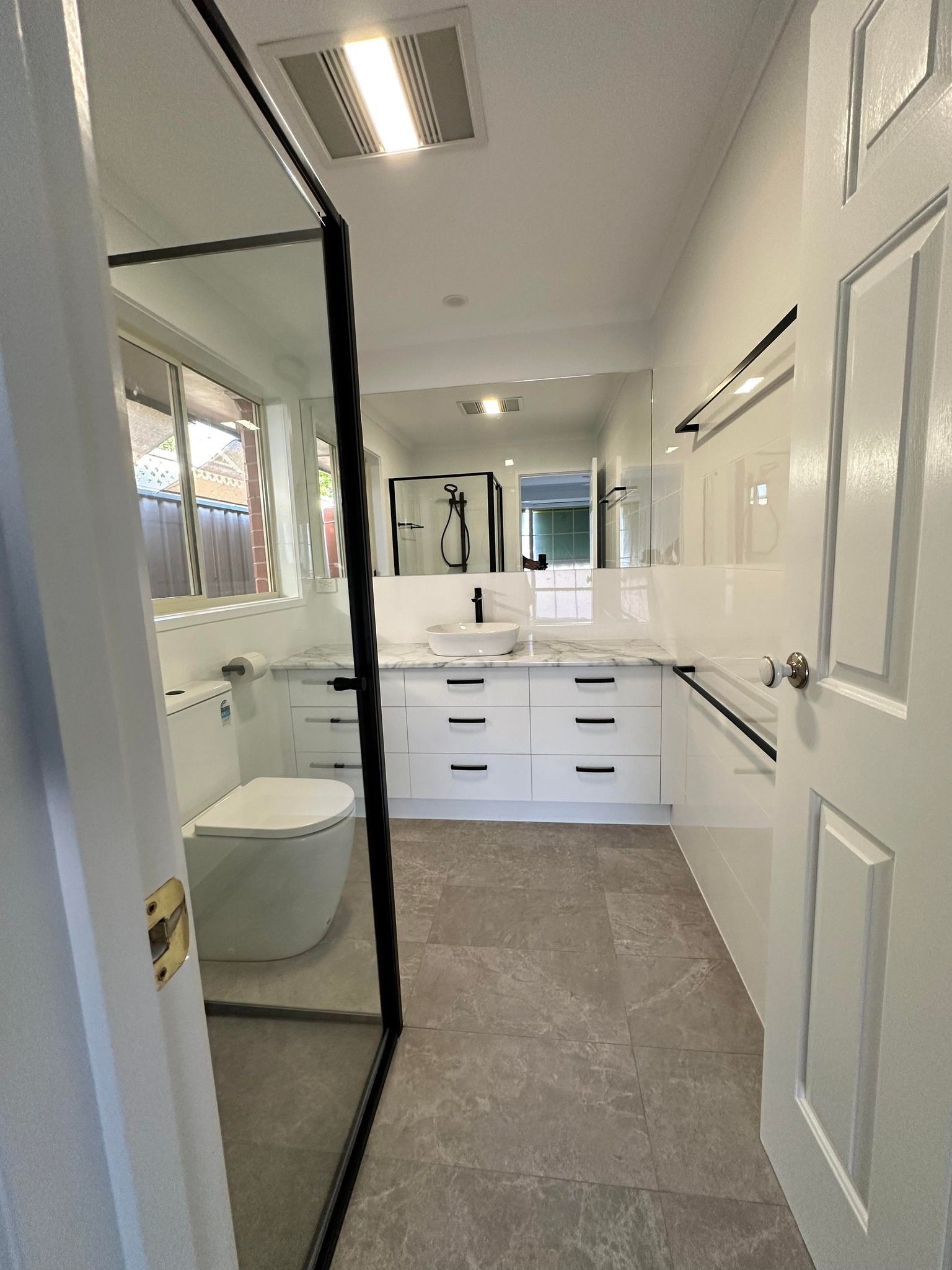 Bathroom with white cabinets, a toilet, a glass shower screen, and grey tile floor.  The space is well-lit and modern.