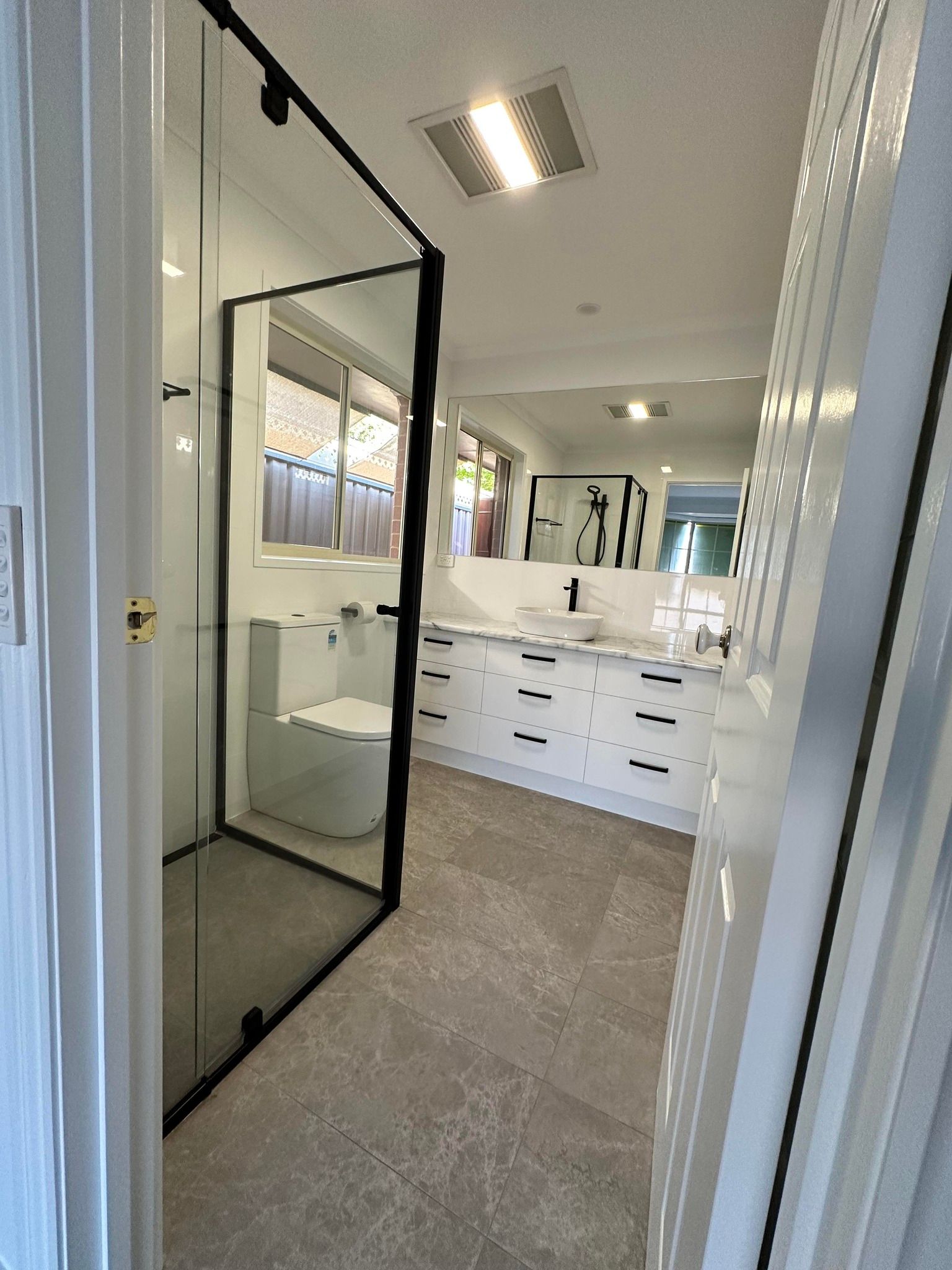 Bathroom with a black-framed glass shower, white vanity with black hardware, and marble-look flooring. The room is bright with a long mirror and a recessed light.