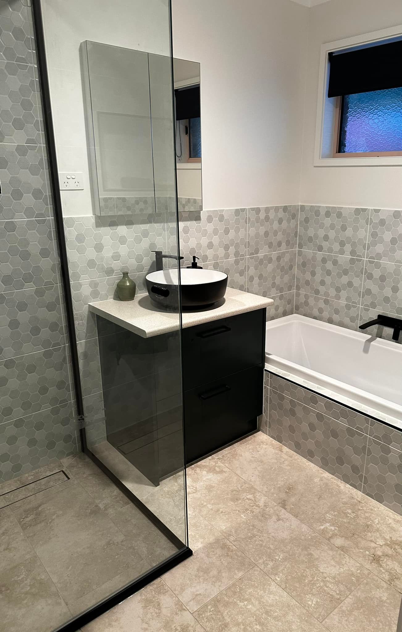 Bathroom with black vanity, white countertop, and black sink. The shower has a glass door, and the walls are tiled in a mosaic pattern.