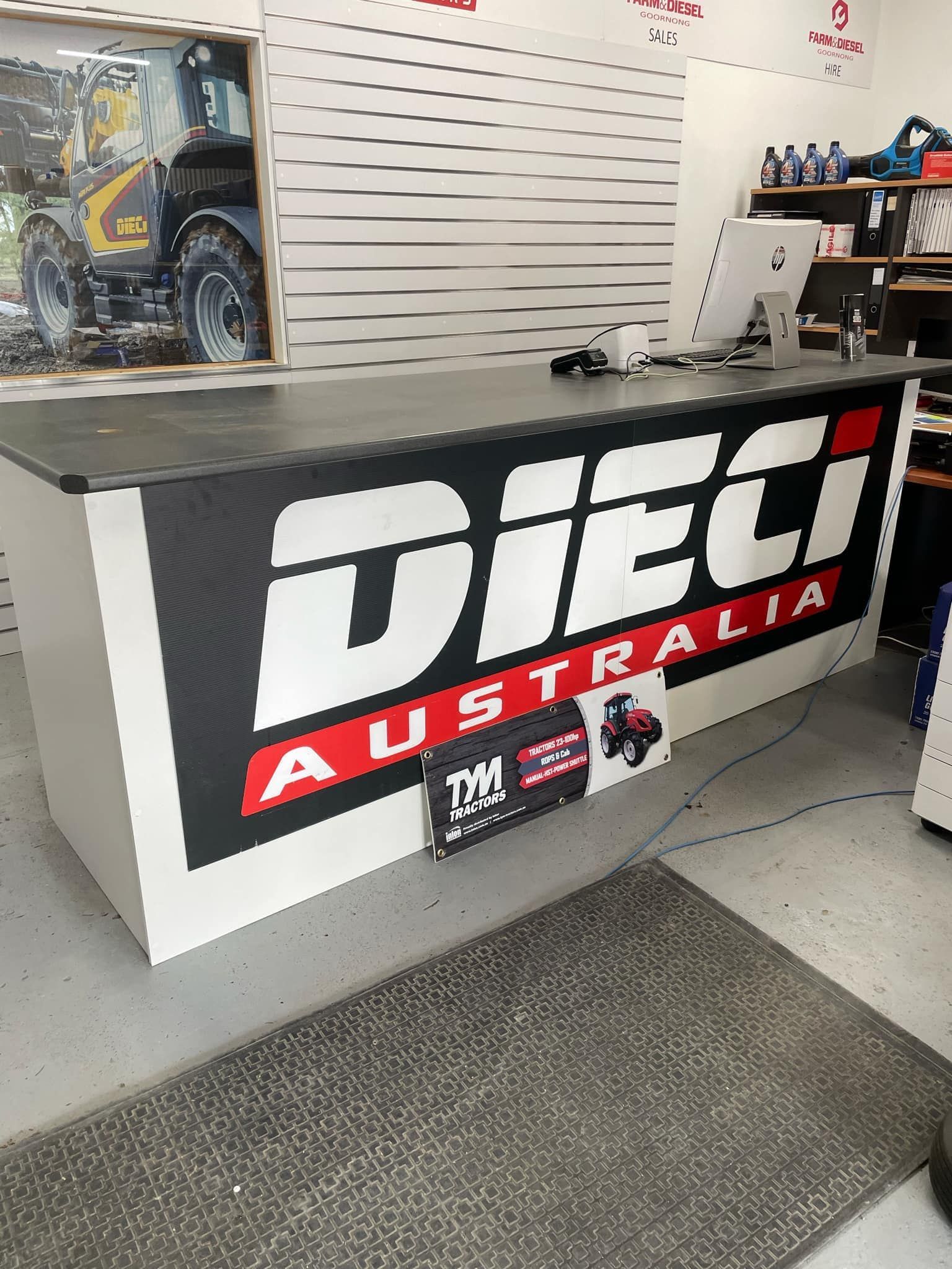 A reception desk with the Dieci Australia logo, set in a workshop environment. The desk is white with black and red branding.