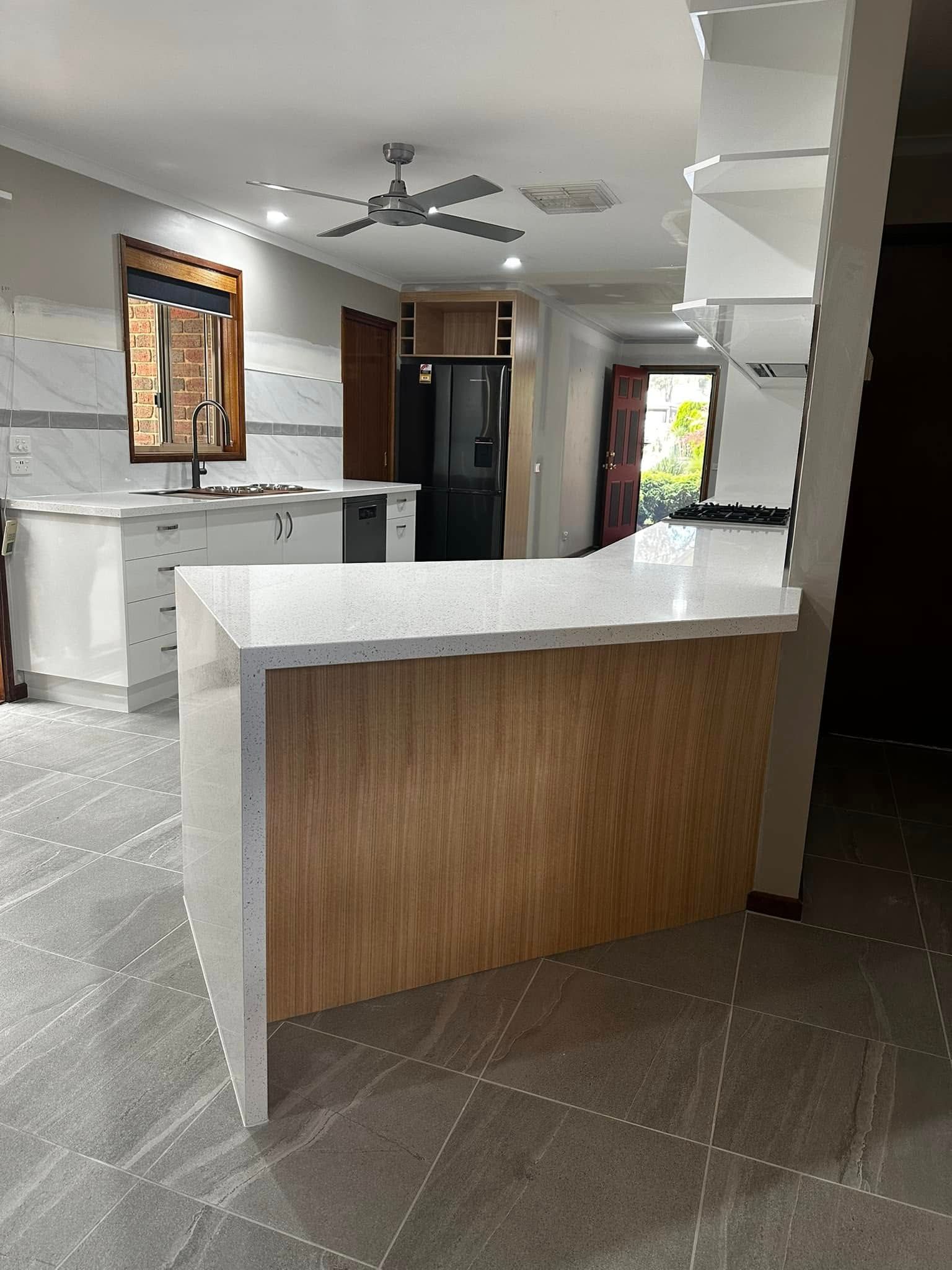 Kitchen with a light wood island, white countertops, and gray tile flooring. A black refrigerator and a view to the outdoors are visible.